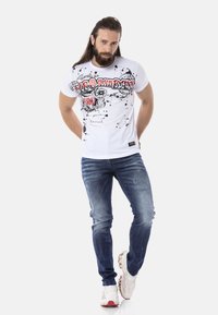 T-shirt blanc à manches courtes avec des impressions graphiques en noir et rouge, associé à un jean bleu déchiré et des chaussures de sport blanches.