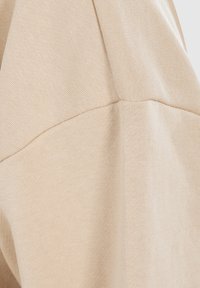 Gros plan sur un tissu beige montrant la texture tricotée détaillée et les lignes de coutures d’un vêtement, probablement un pull ou un sweat.