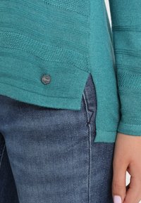 Teal sweater with textured patterns and a side slit, featuring a silver logo button. Paired with dark denim jeans.