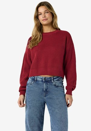 Joven con cabello largo y rubio, vestida con un suéter rojo de punto y jeans claros de talle alto, sobre un fondo blanco.