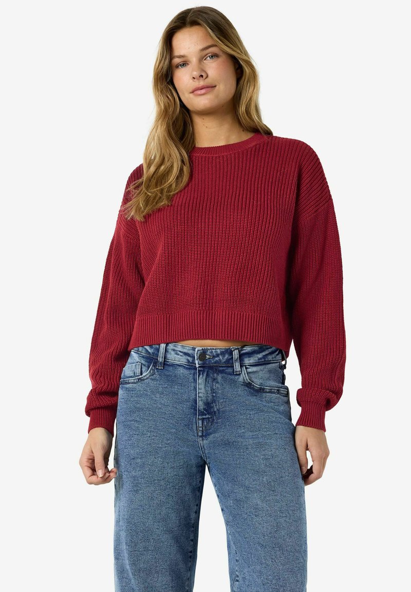Jeune femme aux longs cheveux blonds portant un pull rouge en tricot et un jean taille haute bleu clair sur fond blanc.