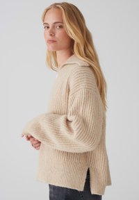 Maglione beige lavorato a maglia con una texture a coste, vestibilità ampia e spacco laterale. Presenta un collo con revers e maniche larghe e oversize.