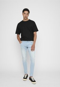 Jeune homme debout devant un fond blanc, portant un t-shirt noir, un jean slim bleu clair et des baskets noires à semelles blanches.