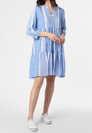 Kobieta ubrana w luźną, niebiesko-białą sukienkę o długości do kolan z rękawami 3/4 i białe sneakersy, stojąca na jednolitym tle.