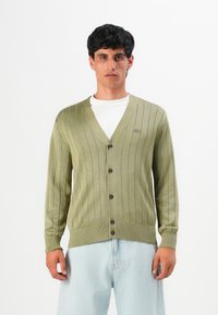 Homem a usar um cardigã canelado verde claro sobre uma camisa branca e jeans azuis claros, de pé contra um fundo branco liso.