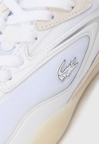 Sneaker bianca con tomaia in rete testurizzata, dettagli in suede, lacci tondi e logo Lacoste argentato sul lato. Suola in gomma con lieve contrasto di colore.