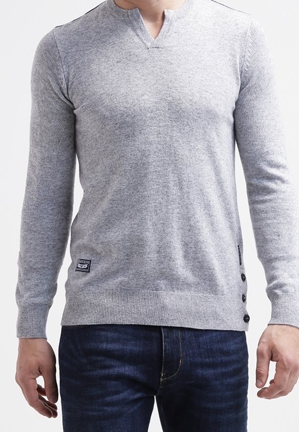 Homme portant un pull gris clair à manches longues avec des boutons sur le côté et un jean bleu foncé, debout devant un fond blanc.