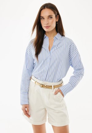 Vrouw met een blauw-wit gestreept overhemd met knopen, ingestopt in witte shorts met hoge taille en een glanzende gouden riem, hand in de zak.