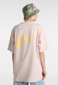 Hellrosa übergroßes T-Shirt mit einem großen gelben "Vans"-Logo auf dem Rücken. Es hat kurze Ärmel und eine lockere Passform. Ergänzt durch einen camouflagen Bucket Hat.