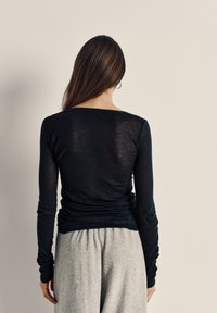 Žena s dugom smeđom kosom u crnoj majici dugih rukava i sivim hlačama, stoji okrenuta prema običnom bež zidu.