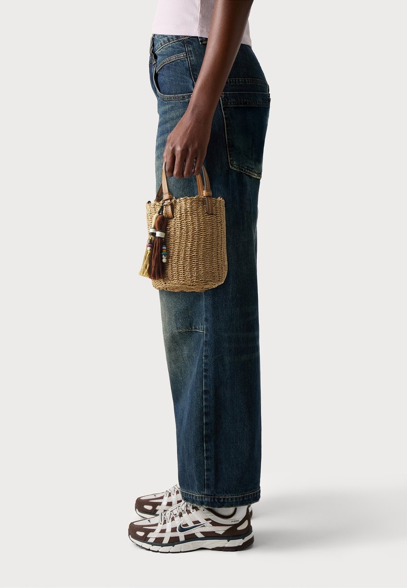 Borsa di paglia intrecciata con manici in pelle, decorata con perline colorate e un pon-pon. Abbinata a jeans blu larghi e sneakers marroni con motivo.