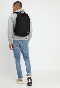 Zaino nero con molte tasche, realizzato in tessuto resistente. Presenta un logo grigio, spallacci regolabili e una forma elegante. Indossato sulla schiena.