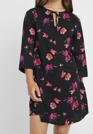 Femme portant une robe noire avec un imprimé floral rose et violet, manches trois-quarts et un petit lien en forme de clé au niveau de l'encolure.