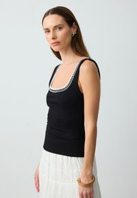 Canotta nera a coste con un ampio scollo bianco a scallop, abbinata a una gonna bianca plissettata e un bracciale dorato, che mette in evidenza una silhouette aderente.