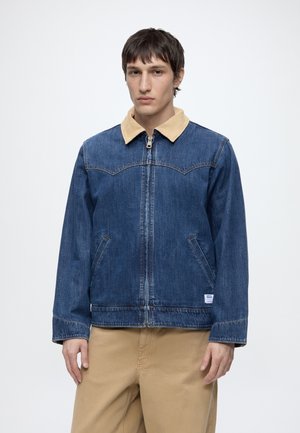 Tânăr purtând o jachetă din denim albastru cu guler bej și pantaloni bej, stând în fața unui fundal alb simplu.