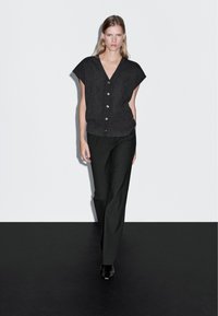 Frau, die in einer ärmellosen schwarzen Knopfjacke, schwarzen Hosen und schwarzen Schuhen vor einem weiß-schwarzen Hintergrund geht.