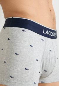 Boxers gris avec ceinture bleu marine arborant "LACOSTE," décorés de petits logos de crocodile bleu marine sur l'ensemble. Composition en mélange de coton, design ajusté.