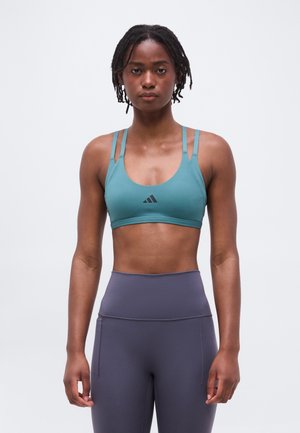 Žena s krátkymi dredmi, oblečená v tyrkysovej športovej podprsenke s dvojitými ramienkami a sivých legínach s vysokým pásom, stojaca pred jednoduchým sivým pozadím.