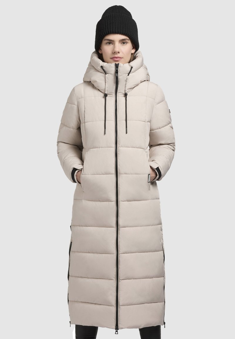Beige long puffer coat with horizontal baffle design, front zipper, drawstring hood, and side zip pockets. Worn with a black knit beanie.