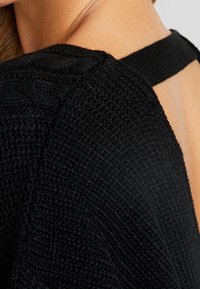 Schwarzes Stricktop mit strukturiertem Design und einem offenen Rücken mit einem dünnen Träger. Der Stoff wirkt weich und dehnbar.