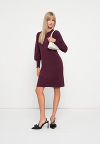 Burgundi kötött ruha hosszú puffujjakkal, térd hosszúságú. Fekete slingback magassarkú cipővel kombinálva, amelyen masni részlet található, valamint egy fehér táskával.
