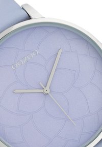 Montre ronde avec un cadran texturé lavande arborant un motif floral, un matériel en métal argenté et un bracelet en cuir bleu clair. Nom de la marque "OOZOO" visible.