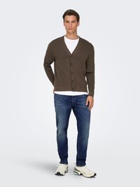 Cardigan en laine marron avec col en V et fermeture boutonnée, porté par-dessus un t-shirt blanc. Assorti à un jean bleu et des baskets claires.