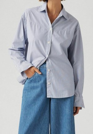 Femme portant une chemise à manches longues rayée bleu clair et blanc, rentrée dans un jean bleu taille haute à jambes larges, avec une main dans la poche.