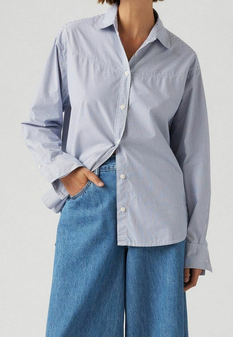 Femme portant une chemise à manches longues rayée bleu clair et blanc, rentrée dans un jean bleu taille haute à jambes larges, avec une main dans la poche.