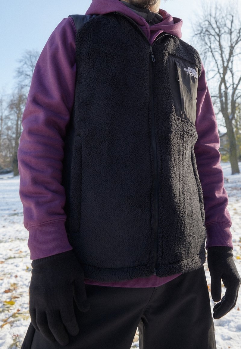 Personne portant un gilet polaire noir, un sweat à capuche violet et des gants noirs, debout à l'extérieur sur un sol enneigé avec des arbres en arrière-plan.