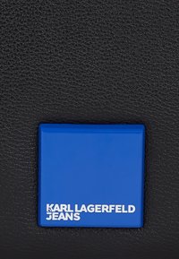 Črn teksturiran material z modrim pravokotnim oznako, na kateri je v belem besedilu "KARL LAGERFELD JEANS". Preprost in moden dizajn.