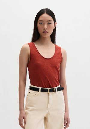 Jeune femme aux longs cheveux noirs portant un haut sans manches couleur rouille rentré dans un pantalon beige taille haute avec une ceinture noire.