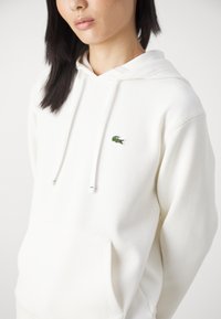 Felpa bianca in cotone, con tasca frontale e coulisse regolabili. Logo del coccodrillo ricamato di colore verde sul petto.
