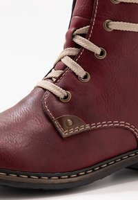 Røde læderankelstøvler med beige snørebånd, metaløjer og brune accenter. Har en struktureret overflade og kontrasterende syninger langs sålen.