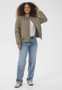 Veste bomber matelassée verte olive avec boutons pression, poignets et ceinture élastiques, associée à un jean bleu à coupe décontractée et des baskets beiges.
