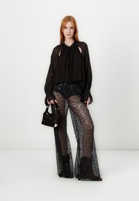 Zwarte transparante blouse met uitsparingen, gecombineerd met zwarte paillettenbroek met een gestructureerd patroon. Het model houdt een kleine zwarte handtas vast.