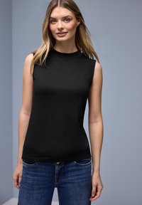 Jeune femme aux longs cheveux blonds portant un haut noir sans manches et un jean bleu, debout devant un fond gris clair uni.