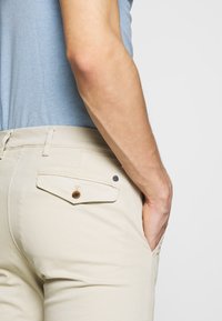 Beige broek met een achterzak met knoopsluiting en een klein zwart accentknopje. Gladde stof met een aangesloten design.