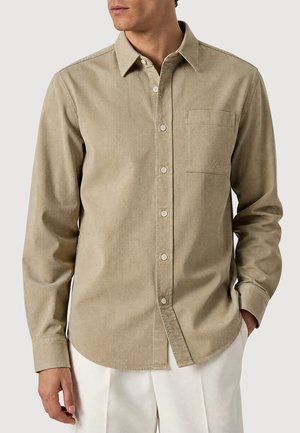 Uomo che indossa una camicia beige con bottoni e taschino sul petto e pantaloni bianchi, in piedi con una mano in tasca su uno sfondo neutro.