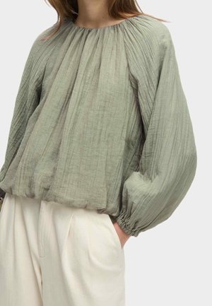 Mujer con una blusa suelta de manga larga y textura verde claro combinada con pantalones de talle alto en color crema con las manos en los bolsillos.