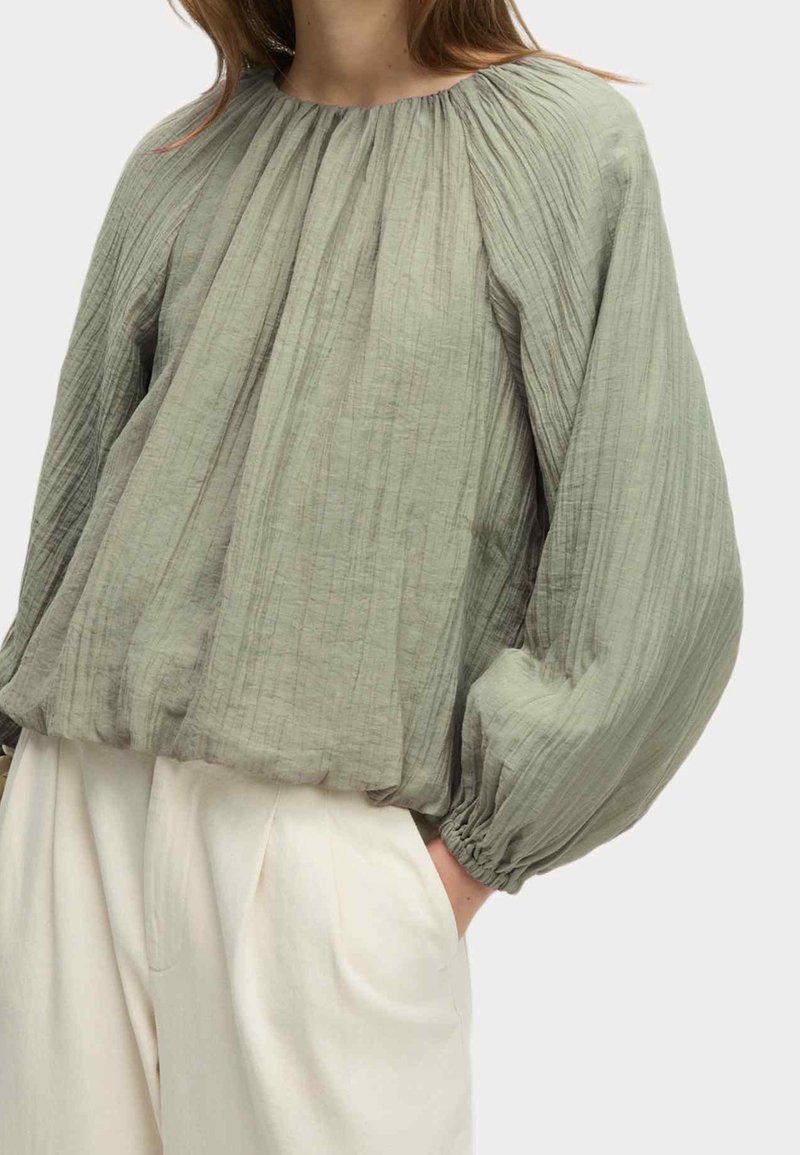 Femme portant un chemisier ample à manches longues texturé vert clair associé à un pantalon taille haute de couleur crème, mains dans les poches.