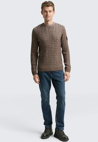 Brauner gestrickter Pullover mit einem strukturierten Muster, rundem Hals und gerippten Bündchen. Kombiniert mit blauen Jeans und schwarzen Stiefeln.