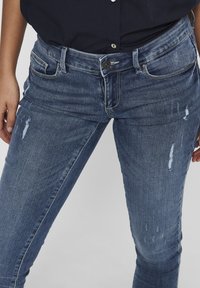 Jeans de mezclilla azul oscuro con un corte ajustado. Presentan detalles desgastados, cinco bolsillos y cierre de botón. Ligeramente desteñidos en los muslos.