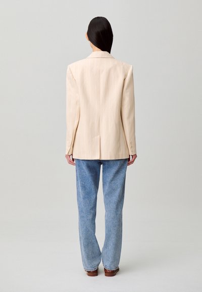 Blazer beige a righe sottili con silhouette strutturata, spacco posteriore singolo e polsini con bottoni, abbinato a jeans azzurri a gamba dritta.