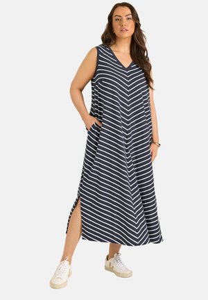 Kvinde står med den ene hånd i lommen, iført ærmeløs marineblå kjole med hvide diagonale striber og sideslids, parret med hvide sneakers.