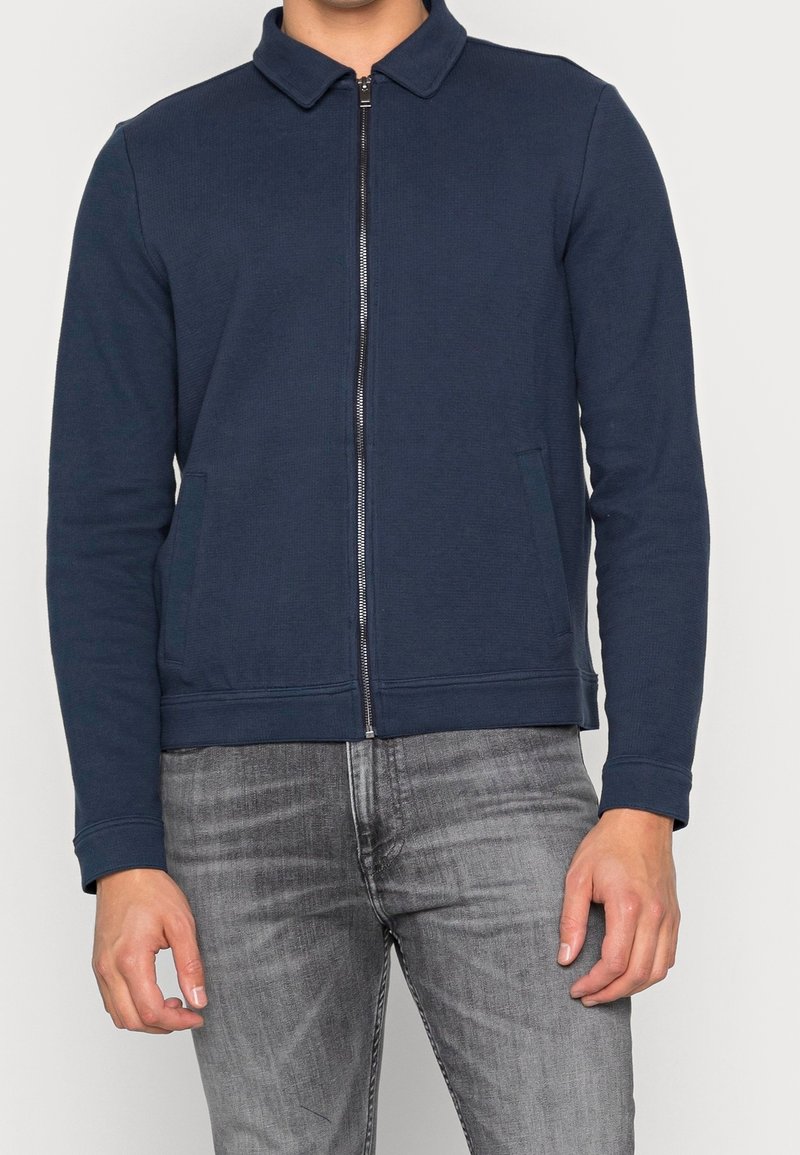 Laivastonsininen vetoketjullinen takki, jossa on kaulus, kaksi sivutaskua, joustavat hihansuut ja sileä pinta. Yhdistetty harmaisiin farkkuihin.