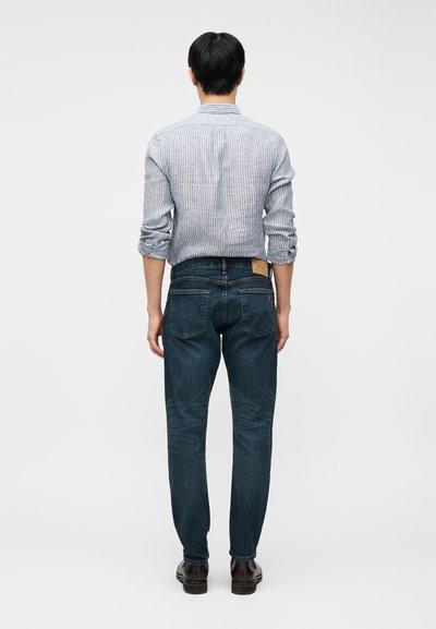 Άνδρας με ριγέ πουκάμισο με τα μανίκια ανεβασμένα, σκούρο μπλε τζιν και καφέ παπούτσια, στέκεται γυρισμένος με την πλάτη σε απλό γκρι φόντο.