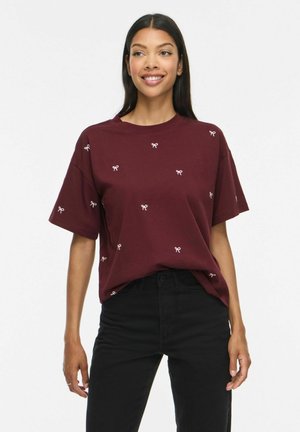 Junge Frau trägt ein bordeauxrotes kurzärmeliges Shirt mit kleinen weißen Schleifenmustern und schwarze Hose mit hohem Bund, lächelt und steht.