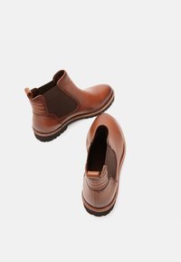 Ruskeat nahkaiset nilkkurit, joissa on joustavat sivut, teksturoitu korko ja kontrastinsaumaus. Pyöreä varvas ja kumipohja pitävyyttä varten.