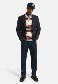 Marineblazer over een gestreepte polo in rood, geel en blauw; gecombineerd met donkere jeans en zwarte instappers, aangevuld met een pet.